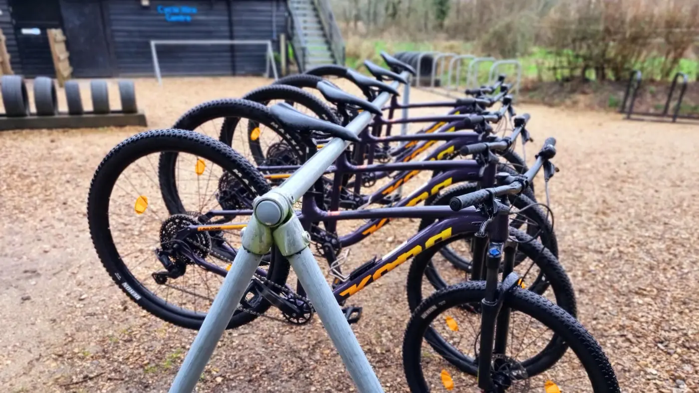 A row of hire bicycles are parked in an outdoor bike rack on a gravel surface at Moors Valley Cycle Hire Centre.