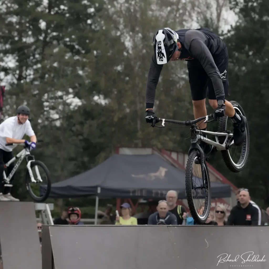 Danny Butler performs a trick mid-air over a ramp while another rider waits on an adjacent ramp. A crowd watches in the background at Pedals at the Park Moors Valley.