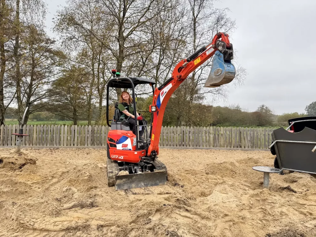 A person operates a small red digger, digging sand on a building site with a wooden fence and trees in the background under a cloudy sky.