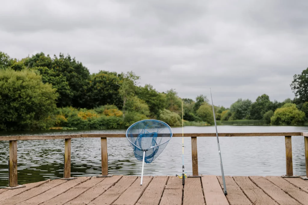 Fishing net and rod placed on a wooden fishing platform overlooking a Moors Lake with greenery and trees in the background under a cloudy sky.