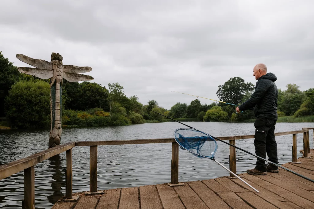 An man stands on a wooden fishing platform by Moors Lake holding a fishing rod. A large dragonfly sculpture is mounted on the dock. A fishing net and more rods rest beside him.