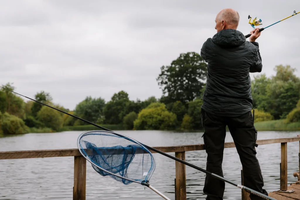 A person in a dark jacket stands on a fishing platform, casting a fishing line into the water, with a blue fishing net beside them.