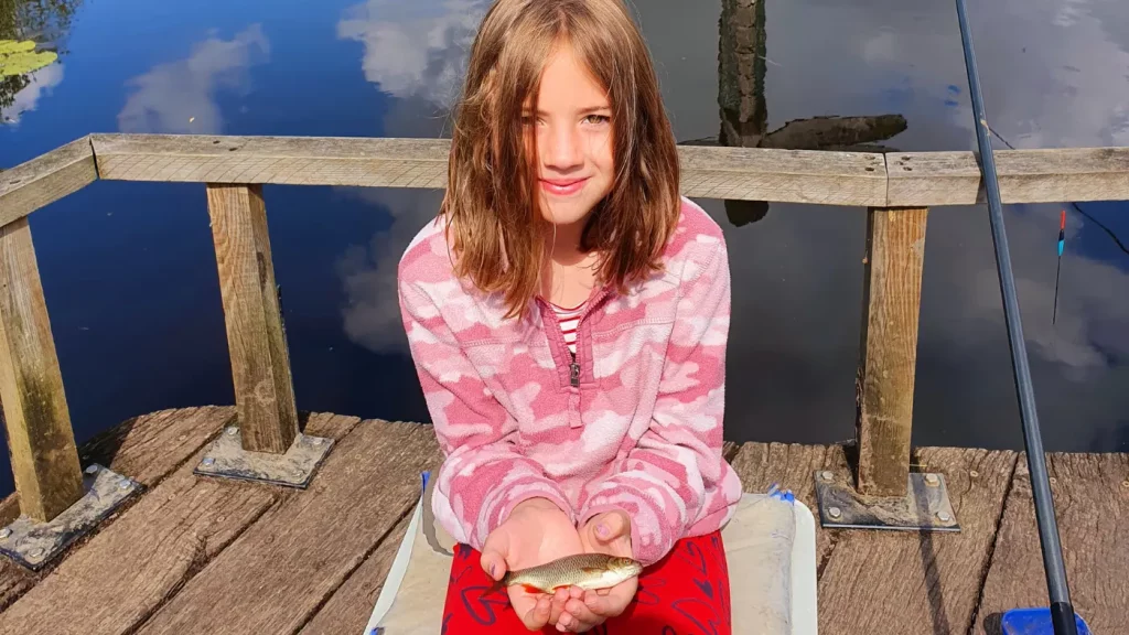 A girl with shoulder-length hair sits on the fishing platform at Moors Lake holding a small fish with both hands. She is wearing a pink camouflage hoodie and red trousers. A fishing rod is visible on the right side of the image.