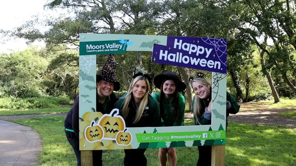 Four rangers in witch hats pose behind a "Happy Halloween" frame at Moors Valley, surrounded by Halloween-themed decorations and greenery.