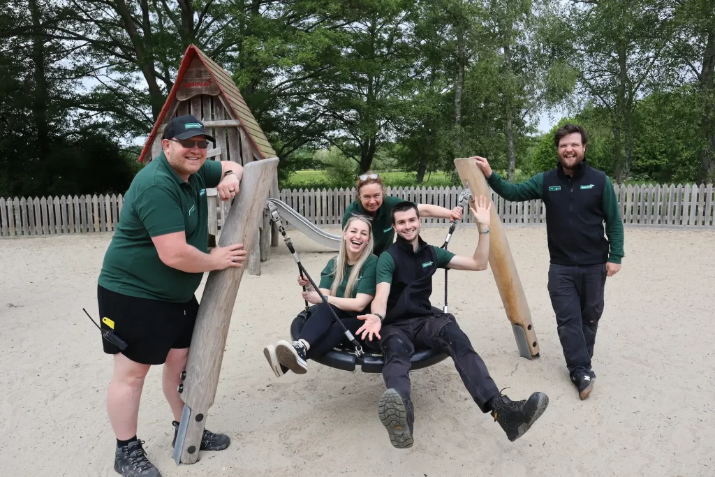 Five smiling rangers in green and black uniforms pose around a wooden swing at a sandy playground, with trees and a wooden fence in the background. Two people sit on the swing whilst others stand nearby.