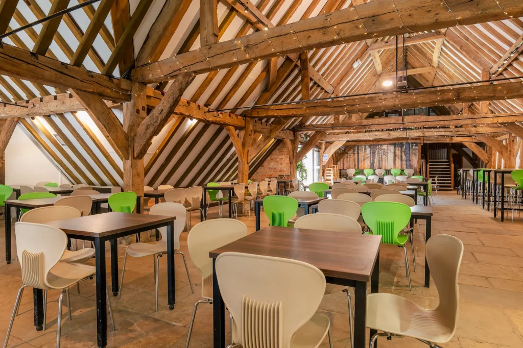 Inside Seasons restaurant showing a spacious dining hall with wooden beams, multiple tables, and chairs. Some chairs are beige, while others are green. The room has a rustic feel with sloped wooden ceilings and stone flooring.