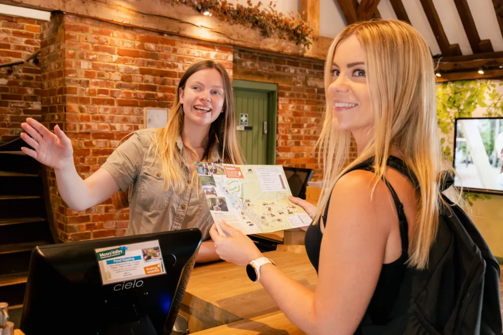 A ranger helps a visitor at the Information Point in the Moors Valley Visitor Centre. The ranger is behind the desk pointing and smiling, while the visitor stands opposite, looking at a map and smiling.