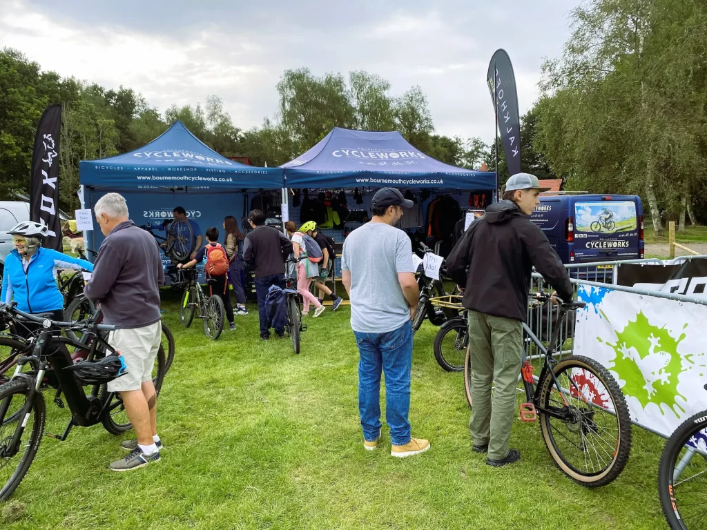 People gather at outdoor stalls displaying bicycles and cycling gear under blue tents with "Cycleworks" branding at Moors Valley Pedals at the Park.