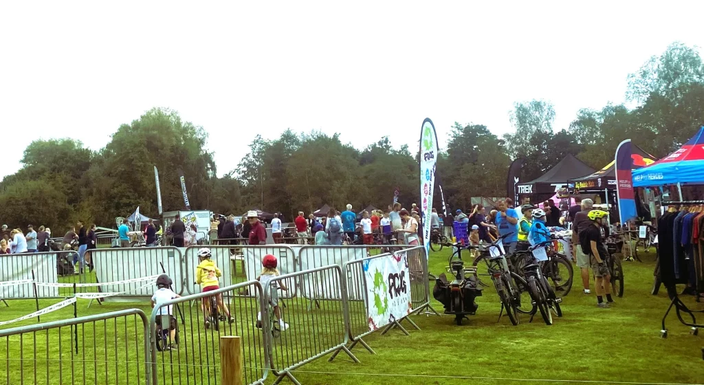 A crowd of people gather at Moors Valley to watch Pedals at the Park, with various vendor booths and bicycles in the foreground.