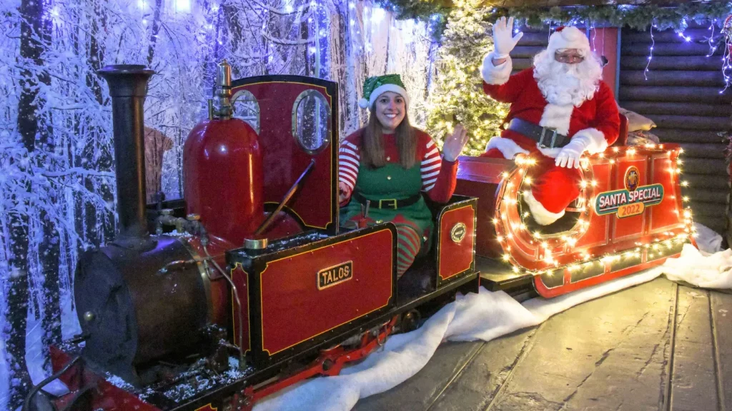 Santa Claus and an elf dressed in festive outfits pose with a decorated miniature train labeled "Santa Special" at Moors Valley Railway, surrounded by Christmas lights and snowy decorations.