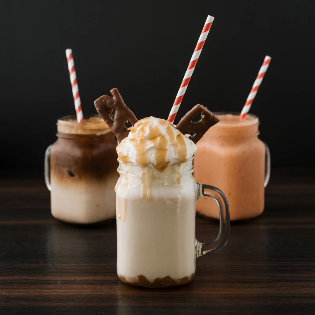 Seasons Drinks. Three mason jar glasses with striped straws, filled with iced coffee, a peach-colored smoothie, and a whipped cream topped shake garnished with caramel syrup and chocolate pieces.