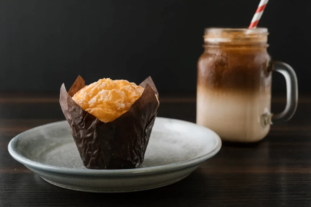 A muffin on a plate and a glass jar with iced coffee topped with foam and a striped straw placed on a dark wooden surface.