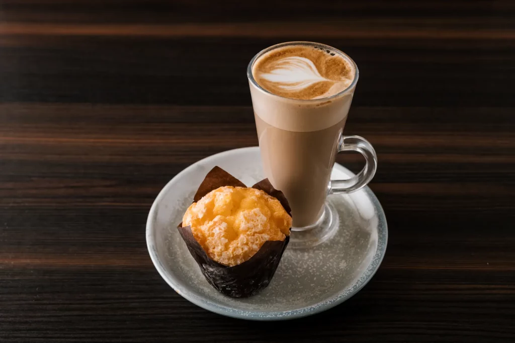 A latte with latte art in a glass mug is placed on a white saucer alongside a wrapped muffin on a dark wooden table.