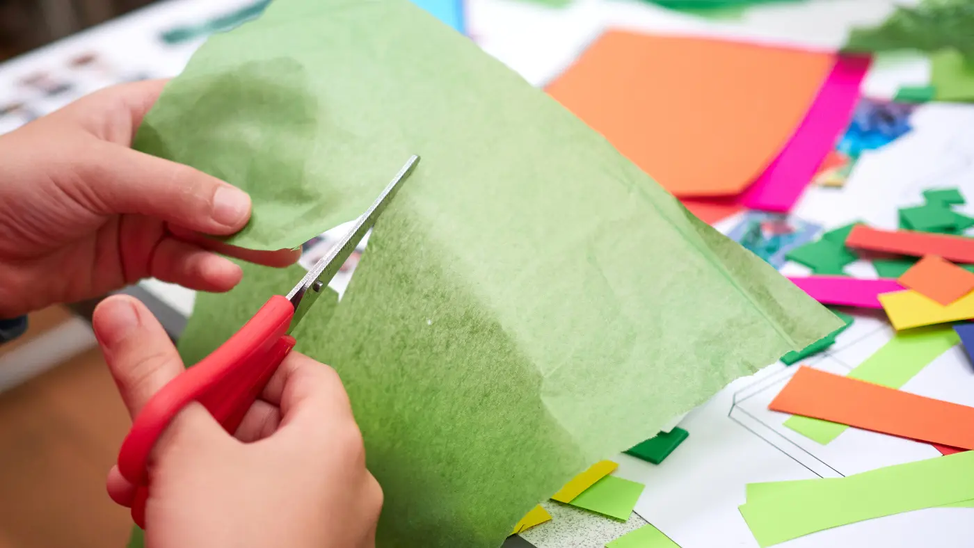 Hands using red scissors to cut a piece of green tissue paper, with colourful paper and craft materials scattered on a table in the background.