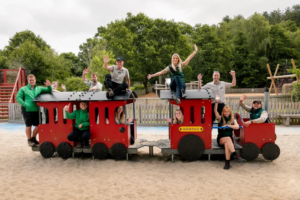 A group of rangers posing and waving on a red and black playground train structure in a sand area, surrounded by greenery and playground equipment in the background.