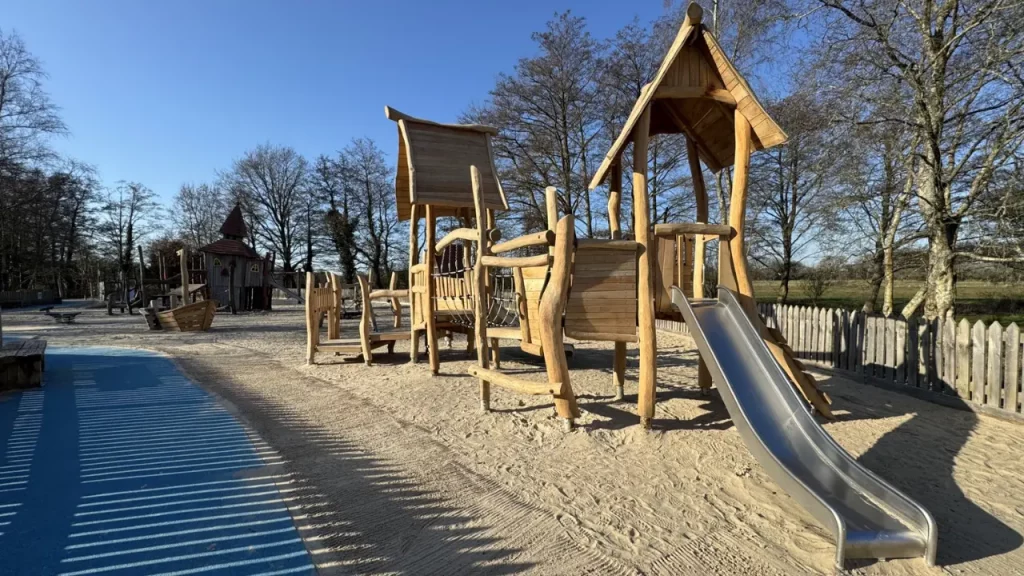 A wooden playground structure with a slide, towers, and rope bridges stands on sandy ground, surrounded by trees and a wooden fence, under a clear blue sky.