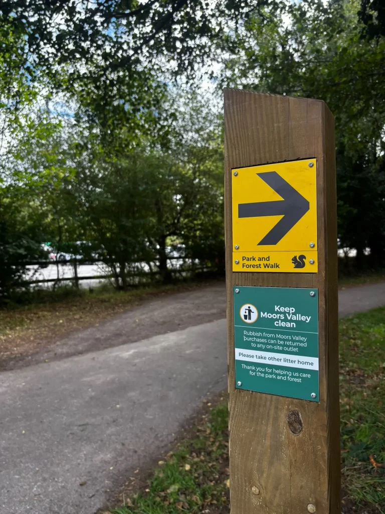 A wooden post with a yellow sign and black arrow points right for the park and forest walk; below is a sign asking visitors to keep Moors Valley tidy by taking rubbish home. Trees and a path are in the background.
