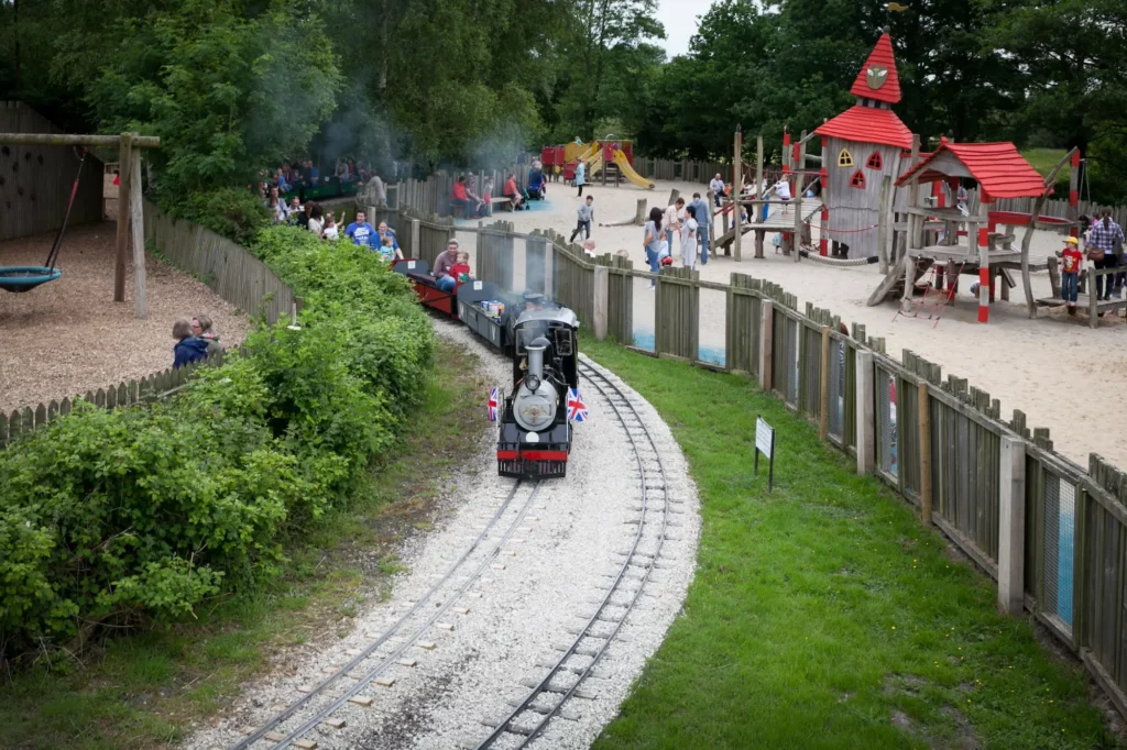 Miniature steam train with passengers on a track in a park, passing a playground with children and adults on a cloudy day.